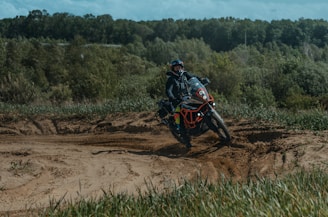 A motorcyclist rides through a sandy path surrounded by green grass and dense forest. The rider is geared up for off-road riding, and dust is being kicked up by the motorcycle tires.