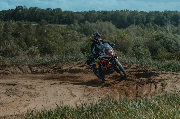 A motorcyclist rides through a sandy path surrounded by green grass and dense forest. The rider is geared up for off-road riding, and dust is being kicked up by the motorcycle tires.