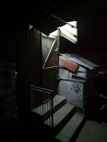 A dimly lit outdoor area with a metal door slightly ajar at the top of a few concrete steps. Corrugated metal sheets form a makeshift roof, casting shadows on the walls. The scene conveys a sense of mystery and isolation.
