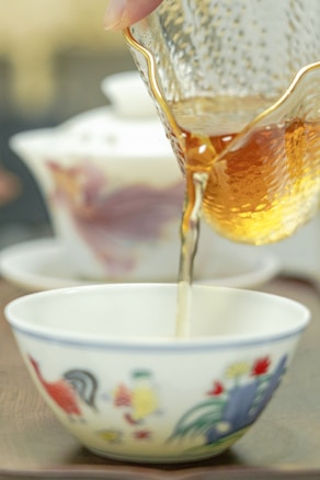 A hand is pouring golden brown tea from a textured glass pitcher into a white ceramic bowl adorned with colorful rooster and floral motifs. In the background, a teapot with similar design elements is visible.