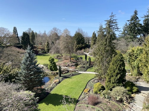A beautifully landscaped garden featuring a variety of trees and bushes. The garden has a well-maintained lawn and a small pond. A winding stone path meanders through the greenery, with vibrant trees and neatly trimmed plants.