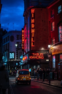 A vibrant street scene at night featuring a neon-lit building with signs for 'Windmill' and 'Tables & Dancing'. People are gathered outside, and a classic yellow car is driving down the street. Other neon signs for a cocktail bar are visible in the background, creating an urban nightlife atmosphere.