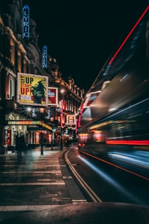 A nighttime urban street scene with illuminated theater signs and marquees. A blur of a passing bus is visible on the right, suggesting motion. The prominent theater signs include names like Lyric and Apollo, and there are posters advertising musicals. The buildings are adorned with various lights, contributing to a vibrant atmosphere.