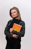A person with medium-length hair is holding an orange book with both hands. The person is wearing a black and white striped shirt over a black top, and is posing confidently against a plain background.