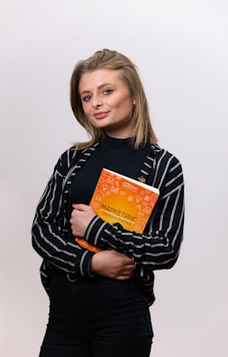 A person with medium-length hair is holding an orange book with both hands. The person is wearing a black and white striped shirt over a black top, and is posing confidently against a plain background.