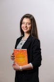 A person stands holding an orange book with white text, wearing a black blazer over a striped shirt. The background is plain and light-colored.