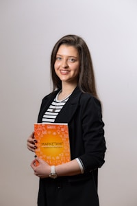A person stands holding an orange book with white text, wearing a black blazer over a striped shirt. The background is plain and light-colored.