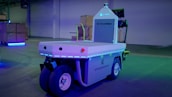A warehouse setting features a modern automated vehicle labeled 'Stockchaser' with significant branding from Columbia and Cynch. The vehicle, equipped with several wheels, shows a futuristic design illuminated by blue LED lights. Surrounding the vehicle, stacks of boxes indicative of a storage or logistics environment add to the industrial atmosphere, and the background is lit with ambient blue and green lights.