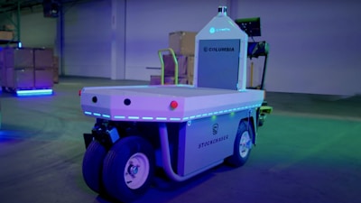 A warehouse setting features a modern automated vehicle labeled 'Stockchaser' with significant branding from Columbia and Cynch. The vehicle, equipped with several wheels, shows a futuristic design illuminated by blue LED lights. Surrounding the vehicle, stacks of boxes indicative of a storage or logistics environment add to the industrial atmosphere, and the background is lit with ambient blue and green lights.