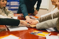 Several people gather around a table placing their hands on top of each other, symbolizing teamwork or unity. The scene is set in an indoor environment with a focus on collaboration. Documents and open books are visible on the table, suggesting a meeting or group study session.