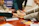 Several people gather around a table placing their hands on top of each other, symbolizing teamwork or unity. The scene is set in an indoor environment with a focus on collaboration. Documents and open books are visible on the table, suggesting a meeting or group study session.