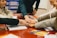 Several people gather around a table placing their hands on top of each other, symbolizing teamwork or unity. The scene is set in an indoor environment with a focus on collaboration. Documents and open books are visible on the table, suggesting a meeting or group study session.