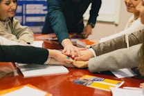 Several people gather around a table placing their hands on top of each other, symbolizing teamwork or unity. The scene is set in an indoor environment with a focus on collaboration. Documents and open books are visible on the table, suggesting a meeting or group study session.