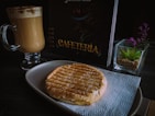 A tabletop setting with a glass mug filled with frothy cappuccino, a grilled sandwich placed on a white napkin on a plate, a menu with the word 'Cafeteria', and a small succulent plant in a square glass pot.
