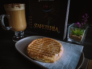 A tabletop setting with a glass mug filled with frothy cappuccino, a grilled sandwich placed on a white napkin on a plate, a menu with the word 'Cafeteria', and a small succulent plant in a square glass pot.