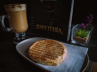 A tabletop setting with a glass mug filled with frothy cappuccino, a grilled sandwich placed on a white napkin on a plate, a menu with the word 'Cafeteria', and a small succulent plant in a square glass pot.