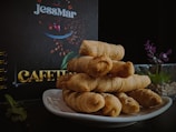 A neatly stacked pile of pastries, possibly breadsticks, is displayed on a white plate against a dark background. In the background, a sign or menu with the word 'CAFETERIA' and the name 'JessMar' is visible. There is a glass container with small plants and some mint leaves on the side.