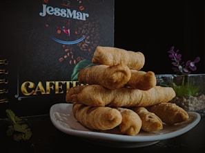 A neatly stacked pile of pastries, possibly breadsticks, is displayed on a white plate against a dark background. In the background, a sign or menu with the word 'CAFETERIA' and the name 'JessMar' is visible. There is a glass container with small plants and some mint leaves on the side.