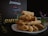 A neatly stacked pile of pastries, possibly breadsticks, is displayed on a white plate against a dark background. In the background, a sign or menu with the word 'CAFETERIA' and the name 'JessMar' is visible. There is a glass container with small plants and some mint leaves on the side.