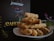 A neatly stacked pile of pastries, possibly breadsticks, is displayed on a white plate against a dark background. In the background, a sign or menu with the word 'CAFETERIA' and the name 'JessMar' is visible. There is a glass container with small plants and some mint leaves on the side.