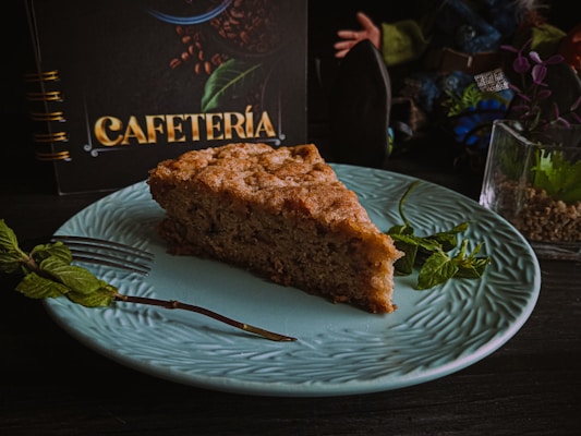 A slice of cake is placed on a textured light blue plate. A sprig of mint lies next to the cake and a silver fork is positioned beside it. In the background, there is a menu or book with the word 'Cafeter&iacute;a' in gold letters, surrounded by coffee bean illustrations, and a small potted plant to the right.