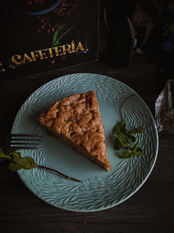 A slice of cake rests on a light blue, textured plate. The cake appears to be a cinnamon or coffee-flavored variety, with a crumbly texture. A sprig of mint is placed next to the cake slice on the plate. In the background, there is a partially visible sign with the word 'CAFETERÍA' in bold yellow letters, and some coffee beans graphics. The overall setting is dark and moody, possibly inside a café or a cozy setting.