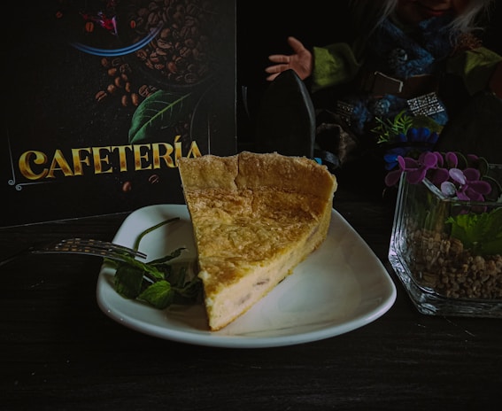 A slice of quiche is placed on a white plate with a metal fork and a few mint leaves. In the background, there is a dark menu with the word 'Cafetería' written in gold, coffee beans, and a green leaf. To the right, a doll with white hair and detailed clothing is partly visible, along with a glass container holding purple flowers and small rocks.