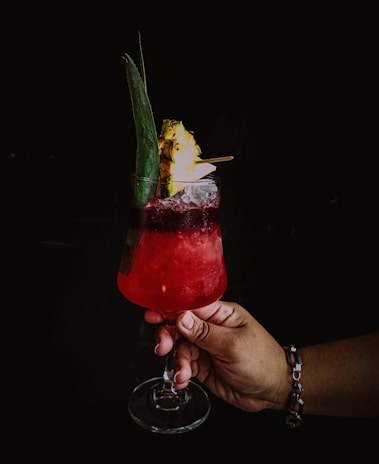 Close-up of a hand holding a cocktail glass filled with a colorful drink, with tropical leaves behind.