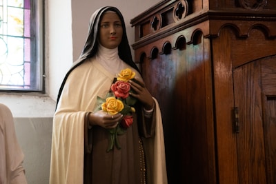 A statue of a woman dressed in a religious habit holding a bouquet of yellow and pink roses, standing next to a wooden confessional booth in a church setting with stained glass windows.
