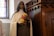 A statue of a woman dressed in a religious habit holding a bouquet of yellow and pink roses, standing next to a wooden confessional booth in a church setting with stained glass windows.