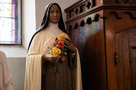 A statue of a woman dressed in a religious habit holding a bouquet of yellow and pink roses, standing next to a wooden confessional booth in a church setting with stained glass windows.