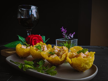 An elegant arrangement of three golden, crispy baskets filled with shrimp and garnished with parsley sits on a plate. A vibrant red rose and a small vase with purple flowers accompany the dish. In the background, a glass of red wine adds a touch of sophistication.