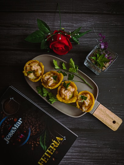 A culinary presentation featuring plantain cups filled with a creamy shrimp mixture is placed on a rectangular dish. Accompanied by fresh parsley sprigs, the setup includes a single red rose and a small, clear glass container with purple and green leaves. A dark wooden table serves as the background, with a menu titled 'JessMar Cafeteria' nearby.