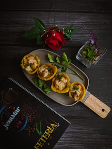 A culinary presentation featuring plantain cups filled with a creamy shrimp mixture is placed on a rectangular dish. Accompanied by fresh parsley sprigs, the setup includes a single red rose and a small, clear glass container with purple and green leaves. A dark wooden table serves as the background, with a menu titled 'JessMar Cafeteria' nearby.