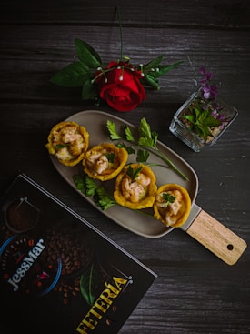 A culinary presentation featuring plantain cups filled with a creamy shrimp mixture is placed on a rectangular dish. Accompanied by fresh parsley sprigs, the setup includes a single red rose and a small, clear glass container with purple and green leaves. A dark wooden table serves as the background, with a menu titled 'JessMar Cafeteria' nearby.
