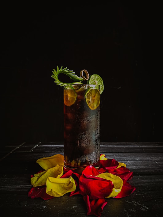 A tall glass of dark, possibly caffeinated, beverage garnished with lime slices and a sprig of greenery. The glass is surrounded by vibrant red and yellow rose petals, all set against a dark, moody background.