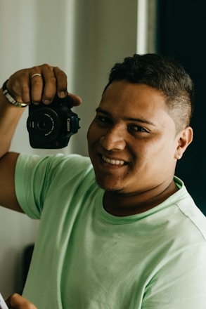 A person is smiling while holding a camera close to their face. They are wearing a light green shirt and have a short haircut. The background is softly blurred, highlighting the subject.