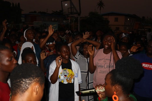 A group of people gathered in an outdoor setting during nighttime. Most individuals have their hands raised, and appear expressive, possibly singing or engaging in a communal activity. A person in the foreground is holding a microphone, suggesting a performance or public speaking event. There are visible buildings and trees in the darkened background, and a basketball hoop is also present.
