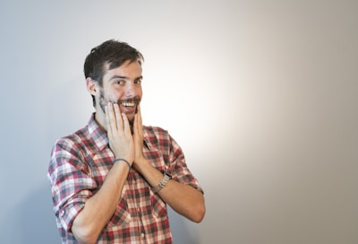 A man with a beard and mustache is wearing a red and white checkered shirt. He has his hands placed on his cheeks and is smiling with an expression that suggests surprise or excitement. The background is plain and light, with a slight shadow cast on one side.
