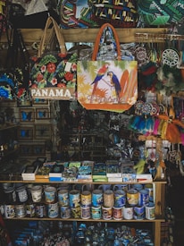 A vibrant souvenir shop display featuring a variety of colorful items including Panama-themed bags, mugs, and decorative objects. The bags showcase designs with tropical birds and floral patterns. Mugs arranged neatly on shelves display various prints, and there are dreamcatchers and other trinkets hanging on the side.