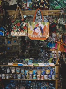 A vibrant souvenir shop display featuring a variety of colorful items including Panama-themed bags, mugs, and decorative objects. The bags showcase designs with tropical birds and floral patterns. Mugs arranged neatly on shelves display various prints, and there are dreamcatchers and other trinkets hanging on the side.