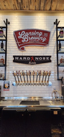 A bar with a white tile backsplash features multiple beer taps lined up in a neat row. Above the taps are signage for Lansing Brewing Company and Hard Nose Spirits. Shelves on either side hold bottles, and a wooden ceiling adds a rustic touch.