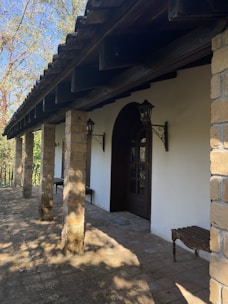 A rustic covered porch with stone pillars and a wooden roof. Two metal lanterns are mounted on the wall beside a wooden door. Benches are placed along the white wall, and the ground is paved with stone tiles.