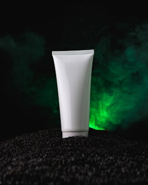 A white squeeze tube stands upright on a mound of black pebbles against a backdrop of swirling green mist. The scene is dramatically lit, highlighting the tube and the mysterious atmosphere around it.
