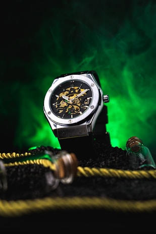 A silver mechanical wristwatch with a visible intricate gear mechanism is placed on a textured surface. The background features a vibrant green light, creating a contrasting effect. Nearby, there are decorative elements like a yellow and black rope and small glass bottles with cork stoppers.