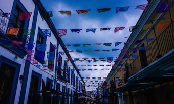 Colorful papel picado banners are strung across a narrow street lined with buildings on either side. The sky is overcast with clouds gathering above, casting a moody tone over the scene. The buildings feature traditional architecture with balconies and brightly colored facades.