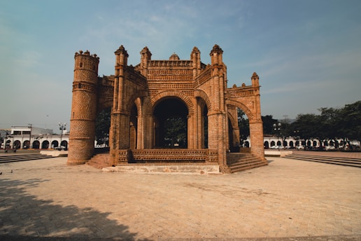 An intricate and historic brick structure with arches and towers stands prominently in the center of a paved plaza. Trees and additional architectural elements can be seen in the background, contributing to a serene and cultural atmosphere.