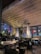 A cozy indoor seating area with dark metal chairs and wooden tables, surrounded by lush green plants. Two tall, pyramid-shaped patio heaters add warmth and ambiance. Outside, colorful lights are visible through large glass windows, enhancing the inviting atmosphere.