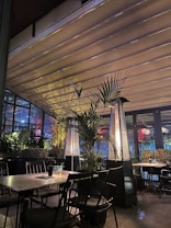 A cozy indoor seating area with dark metal chairs and wooden tables, surrounded by lush green plants. Two tall, pyramid-shaped patio heaters add warmth and ambiance. Outside, colorful lights are visible through large glass windows, enhancing the inviting atmosphere.