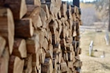 A stack of neatly arranged cut firewood, displayed along the side of a building or structure. The logs are uniformly cut, showcasing the rings of the wood. In the background, an out-of-focus view of a natural landscape with trees and a path can be seen.